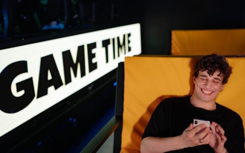 Smiling young man gaming on smartphone in a cozy indoor setting with 'Game Time' sign.
